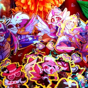 May include: A selection of vibrant cartoon character keyrings featuring winged figures, hearts, and halos. The keyrings showcase characters in pink, purple, and yellow hues. Some keyrings have the text "SunnyCitrineCrafts".