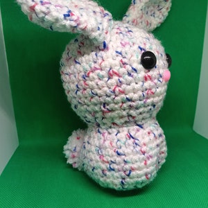 May include: A handmade crochet bunny in white yarn with flecks of blue, pink, and purple. The bunny has large black eyes and a small pink nose. The ears are upright, and the bunny is sitting.