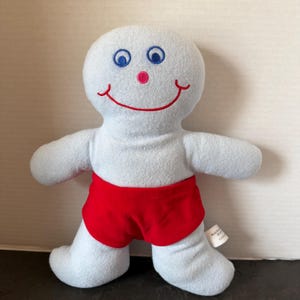 May include: A light blue plush doll with a rounded head and body, featuring embroidered blue eyes, a small pink nose, and a wide red stitched smile. The doll wears bright red fabric shorts and has a small white tag attached to the side.