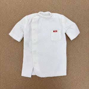 May include: A white doll-sized shirt with short sleeves and a small pocket on the chest. The word "KEN" is embroidered in red on the pocket. The shirt has a button-up front and a rounded collar. The garment is made of a lightweight fabric.
