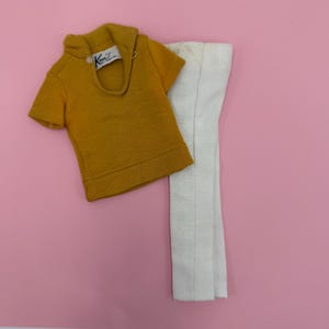 May include: Mustard yellow short-sleeved top with a collar and a white tag. The top is paired with white pants. The outfit is displayed on a pink background. The tag reads "Ken TM Mattel".