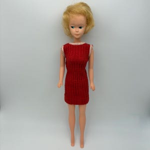 May include: A vintage doll with blonde hair and blue eyes, wearing a red knitted dress. The doll is standing upright against a plain white background. The dress has a textured pattern and a simple, classic design.