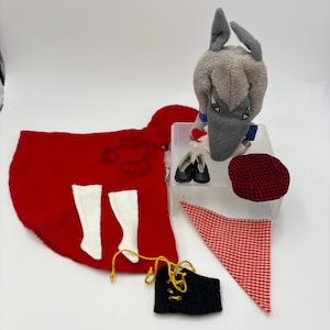 May include: A collection of doll clothing and accessories, including a red cape, white socks, black shoes, a red and black checkered cap, a red and white checkered bandana, and a black vest with yellow laces. A gray wolf doll is also present.