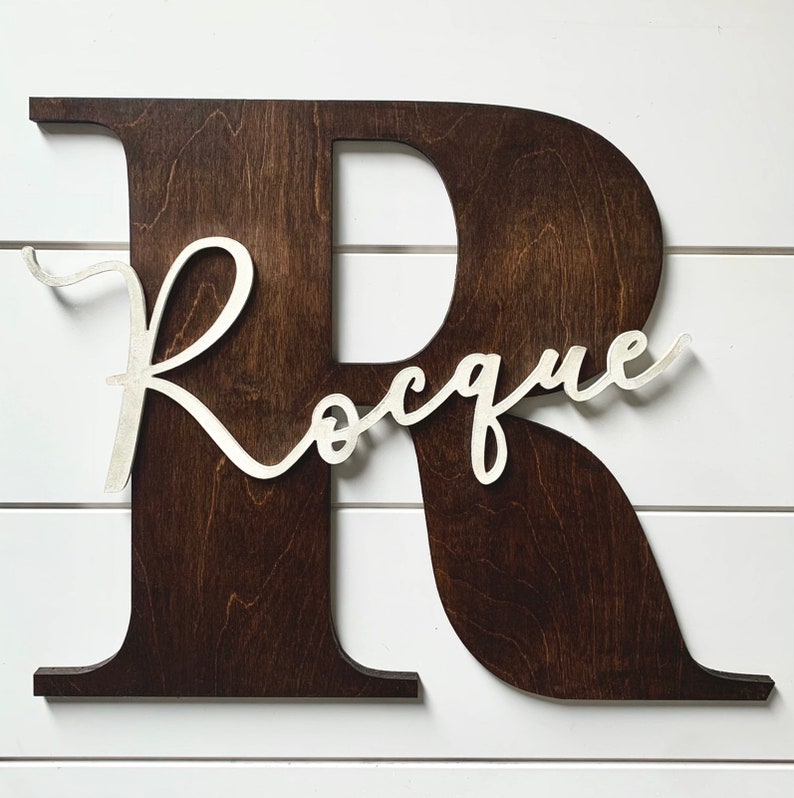 May include: A dark brown wooden letter R with the name "Roque" written in white cursive script.
