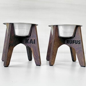 Peut inclure: Deux bols pour chien en bois avec des bols en acier inoxydable. Les bols sont surélevés sur un support en bois à quatre pieds. Le support de gauche est étiqueté "KAI" et le support de droite est étiqueté "RUFUS".