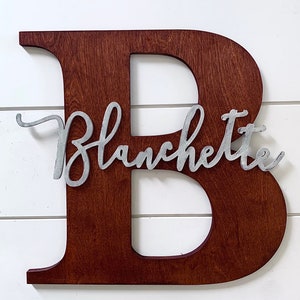 May include: A wooden letter B painted in a dark brown colour with the name "Blanchette" written in silver metal letters on the front of the letter.