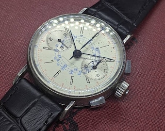 May include: A silver chronograph wristwatch with a white face and black leather strap. The watch has a black second hand, a red minute hand, and a blue hour hand. The watch face has the words "WORD WATCH" and "MAGNETIQUE" printed on it.