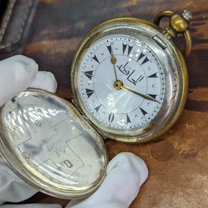 May include: A silver pocket watch with a white face and black Arabic numerals. The watch has a gold-colored case and a gold-colored chain. The watch face has the word "الظفا" written in Arabic script.
