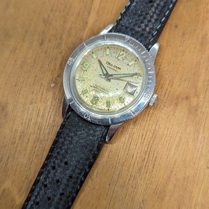 May include: A silver Bulova Endowed Automatic watch with a black leather band. The watch face is cream colored with black markings and a date window at the 3 o'clock position.