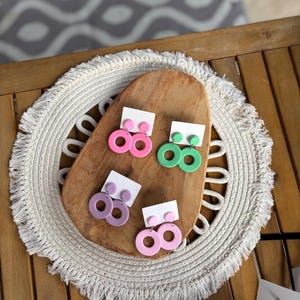 Könnte beinhalten: Vier Paar bunte, runde Ohrringe auf einem Holzbrett präsentiert. Die Ohrringe sind in den Farbtönen Rosa, Grün und Lila gehalten. Sie werden auf kleinen weißen Karten präsentiert, was darauf hindeutet, dass sie zum Verkauf stehen. Der Hintergrund umfasst einen dekorativen Tischset und einen Holztisch.