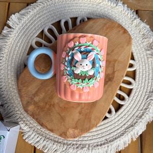 Puede incluir: Una taza de cerámica color melocotón con un asa azul. La taza presenta un diseño decorativo con temática de Pascua con un conejito blanco en una cesta, rodeado de detalles florales rosas y verdes. La taza está colocada sobre una tabla de cortar de madera.