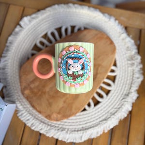 Könnte beinhalten: Eine hellgrüne Keramiktasse mit einem pfirsichfarbenen Henkel. Die Tasse hat ein Ostermotiv mit einem Hasen in einem Korb, umgeben von Blumen. Die Tasse steht auf einem Holzschneidebrett und einem weißen Platzset.