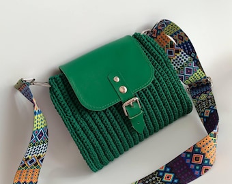 May include: A green crossbody bag with a knitted body and a flap closure. The flap is made of smooth green material and secured with a silver buckle. The bag features a colourful patterned strap.