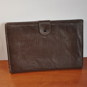 Op de afbeelding: Een bruine leren portfolio met een gestructureerd oppervlak en een klep sluiting met een knoop.