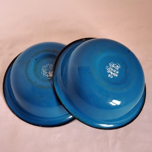 Op de afbeelding: Twee blauwe emaille kommen met zwarte randen. De kommen zijn op elkaar gestapeld. De kommen hebben een wit bloemenmotief op de bodem.