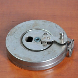 Puede incluir: Una cinta métrica metálica vintage en una caja circular. La cinta métrica es de color plateado con un pequeño asa y un anillo metálico articulado. La caja muestra signos de desgaste, lo que sugiere edad y uso. La cinta métrica está sobre una superficie de madera.