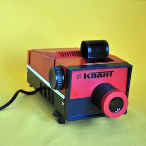 May include: A vintage red and black slide projector with the word "KBAHT" printed on the front. The projector has a black lens and a black power cord. The background is yellow.