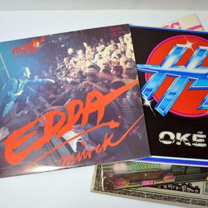Puede incluir: Tres discos de vinilo con diferentes portadas de álbum. El primer disco tiene un diseño rojo y blanco con el texto "EDDA" y "Münich" en él. El segundo disco tiene un diseño azul y plateado con el texto "OKÉ" en él. El tercer disco tiene un diseño rojo y blanco con el texto "rocker" en él.
