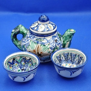 May include: A ceramic tea set with a teapot and two small bowls. The teapot has a blue, green, and white floral design with a matching lid. The bowls have a similar pattern. The set is on a blue background.