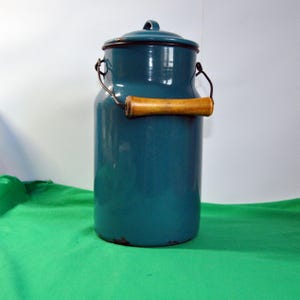 Puede incluir: Una lechera de esmalte azul verdoso con un asa de madera. La lechera tiene una tapa y está hecha de metal.