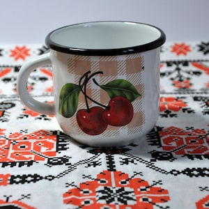 May include: White enamel mug with a black rim and a checkered pattern. The mug features a cherry design with green leaves.