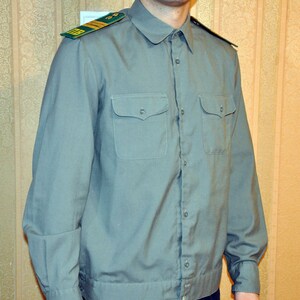 May include: A light blue long-sleeved shirt with a button-up front and two chest pockets. The shirt has a green and gold patch on the left shoulder.