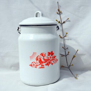 May include: A white enamel milk can with a lid and handle. The can features a red floral and bird design. The rim and handle attachments are black. A sprig of a budding plant is next to the can.