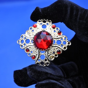 May include: A vintage brooch with a gold-tone filigree design. The brooch features a large, faceted red gemstone at the center, surrounded by smaller red stones and decorative openwork. The brooch is held against a blue background.