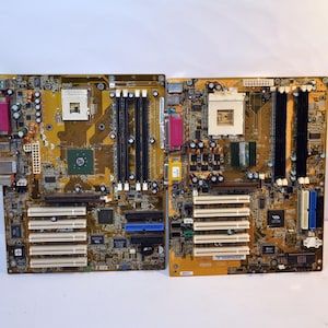 Op de afbeelding: Twee computer moederborden met goud- en bruinkleurige printplaten. Elk bord heeft meerdere slots, poorten en chips. Eén bord heeft een roze poort en de andere een blauwe poort. De borden staan op een witte achtergrond.