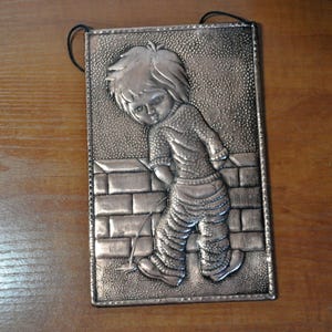 May include: A silver metal wall hanging depicting a child standing in front of a brick wall and urinating. The child is wearing a sweater and pants.