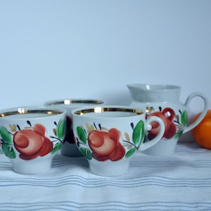 Puede incluir: Un juego de cuatro tazas de té blancas con diseños florales y ribetes dorados, junto con una jarra de crema a juego. Las tazas y la jarra de crema presentan flores pintadas en rojo, verde y dorado. Una sola naranja se encuentra sobre un mantel a rayas blancas y azules.