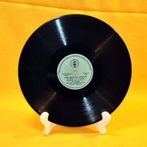 Peut inclure: Un disque vinyle noir avec une étiquette comportant du texte cyrillique et un emblème circulaire. Le disque est présenté sur un support blanc sur fond jaune. L'étiquette comprend le texte "ЗОРІ ЯСНО НАД ДНІПРОМ."