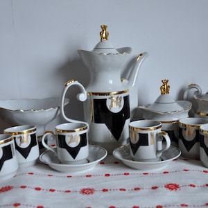 Könnte beinhalten: Ein weißes Porzellan-Tee-Set mit goldenen Akzenten und schwarzen und goldenen geometrischen Mustern. Das Set enthält eine Teekanne, eine Milchkännchen, eine Zuckerdose und sechs Tassen und Untertassen.