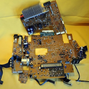 May include: Two brown circuit boards with various electronic components, including capacitors, resistors, and integrated circuits. One board has a large silver heat sink. Black ribbon cables and other connectors are attached.