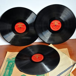 Puede incluir: Tres discos de vinilo negros con etiquetas rojas y doradas. Los discos están colocados sobre una superficie marrón claro, uno de ellos parcialmente cubierto por una funda de papel beige. Las etiquetas presentan texto cirílico y un escudo.