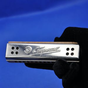 May include: A silver harmonica with a wooden top is held against a blue background. The harmonica has the word "Спутник" in cursive script. There are four holes on each end of the harmonica. The harmonica is held by a person wearing a black glove.