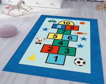 May include: A rectangular children's rug with a hopscotch design. The rug features a light blue background with a hopscotch grid in various colors, including red, yellow, green, and blue. The border is a solid blue. The rug also includes a soccer ball and a teddy bear.