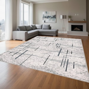 May include: A rectangular rug featuring a cream-coloured base with a geometric design of black and grey lines. The rug is positioned on a wooden floor within a living room, with a grey corner sofa and a fireplace visible in the background.
