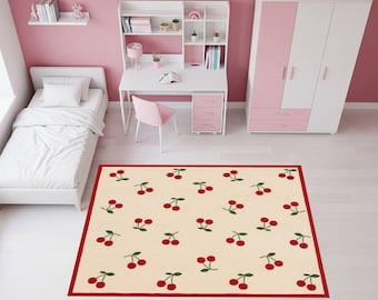 May include: A rectangular rug with a cream-colored background and a repeating pattern of red cherries with green leaves. The rug has a red border and is placed on a white floor in a pink room. The room also features a white desk, bed, and wardrobe.