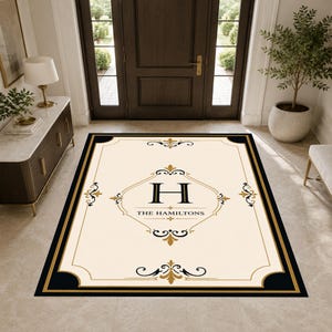 May include: Cream-colored rectangular welcome mat with a black and gold border. The mat features a large black letter "H" inside a gold-lined diamond shape, with "THE HAMILTONS" below. Decorative gold flourishes adorn the corners.