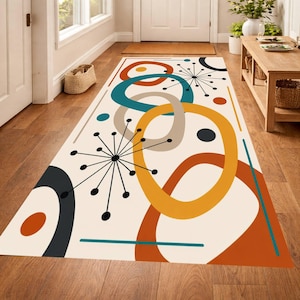 May include: A rectangular area rug with a mid-century modern design. The rug features overlapping circles and starburst patterns in teal, orange, yellow, gray, and black on a cream background. The rug is placed on a wooden floor.