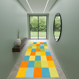 Könnte beinhalten: Ein farbenfroher rechteckiger Teppich mit einem geometrischen Muster aus Quadraten in Gelb, Orange, Blau und Grün. Der Teppich liegt in einem Flur mit hellgrünen Wänden, einem großen Fenster und einem dekorativen Spiegel.