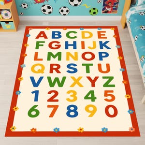 May include: A rectangular area rug featuring a cream-colored center with the alphabet from A to Z and numbers from 1 to 0 printed in various colors including red, blue, green, yellow, and orange, surrounded by a red border with small flower icons.