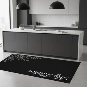 Op de afbeelding: Een zwarte rechthoekige keukenmat met een wit bloemmotief en de woorden "My Kitchen" in cursief. De mat is geplaatst voor een donkergrijs keukeneiland met een wit aanrechtblad en zwarte hanglampen.