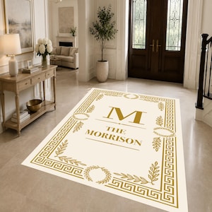 May include: A rectangular area rug with a cream-colored background and a gold-colored border featuring a Greek key pattern and laurel wreaths. The center displays a large gold letter M above the text THE MORRISON in gold lettering.