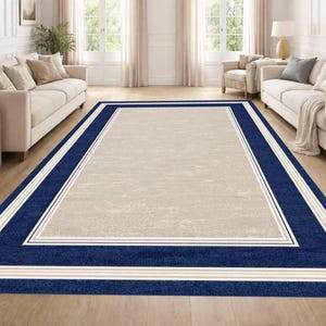 May include: A rectangular rug with a beige central area and a navy blue border. The border has white and beige stripes. The rug is on a wooden floor in a living room, with a light-colored interior.