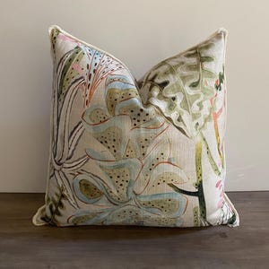 May include: Square decorative pillow with a botanical print. The pillow features a cream-colored background with a colorful floral design in shades of green, blue, pink, and orange. The pillow has a white trim.