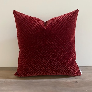May include: A square, burgundy-colored decorative pillow with a textured, geometric pattern. The pillow is made of a soft, plush material and is placed on a wooden surface against a white wall.