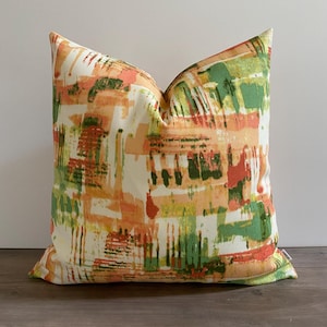 May include: Square decorative pillow with a colorful abstract design. The pillow features a cream-colored background with overlapping brushstrokes in shades of orange, green, and red. The pillow is placed on a wooden surface.