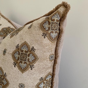 May include: Close-up of two decorative throw pillows. The pillows feature a beige background with a repeating pattern of brown and gold floral designs. The edges are trimmed with a fringed border in a matching brown color. The pillows are a soft, textured fabric.